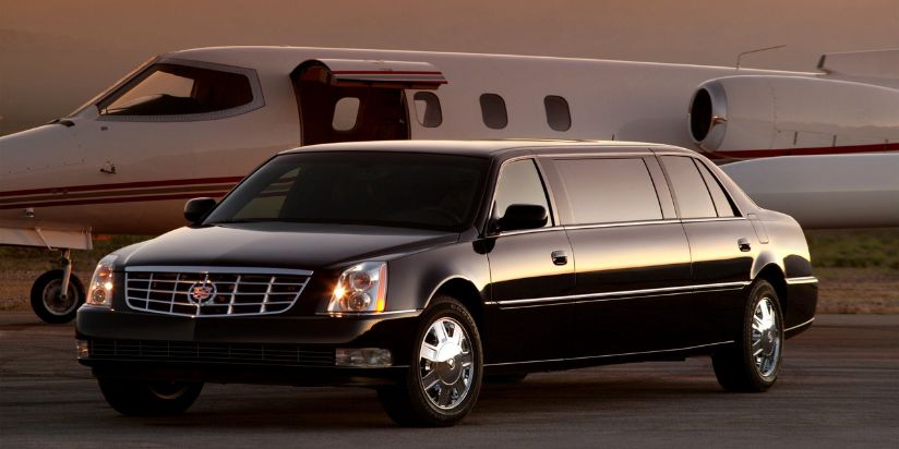 A black limousine is parked at the airport, with a jet parked next to it, and a chauffeur inside the limousine