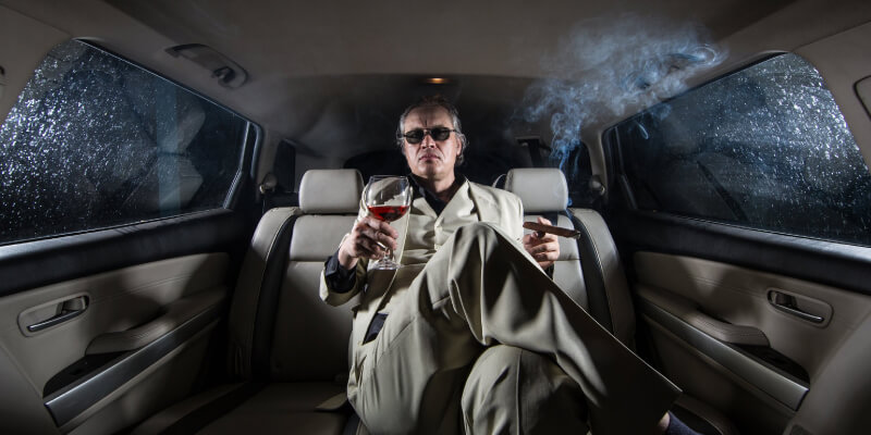 A boss sits in a limousine holding a glass and a cigarette.